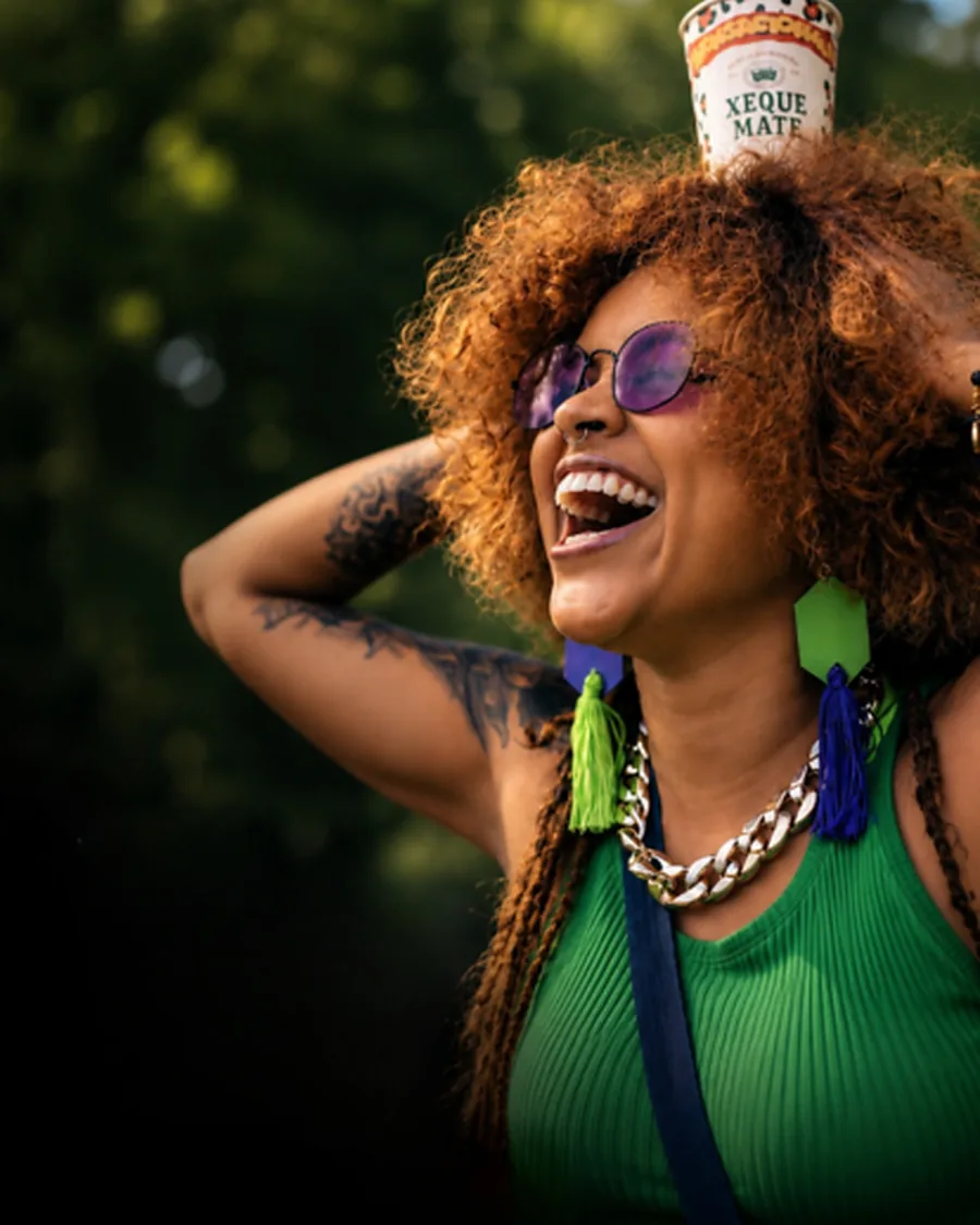 Mulher rindo em um evento ao ar livre com um copo Xeque Mate equilibrado na cabeça